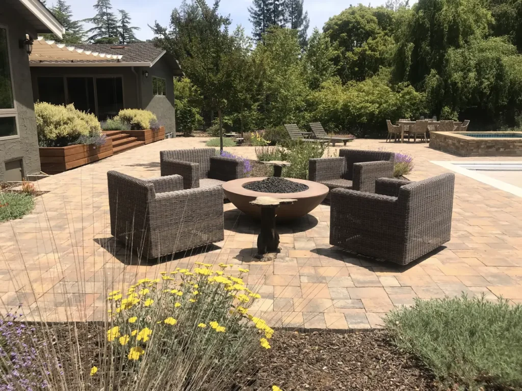 Spacious interlocking paver patio with dark wicker seating around a modern fire pit, bordered by landscaped garden beds and a pool.