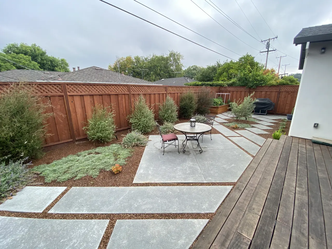 Modern backyard with large geometric concrete pavers, smooth stepping slabs, a wooden deck, paved patio, and landscaped planting beds.