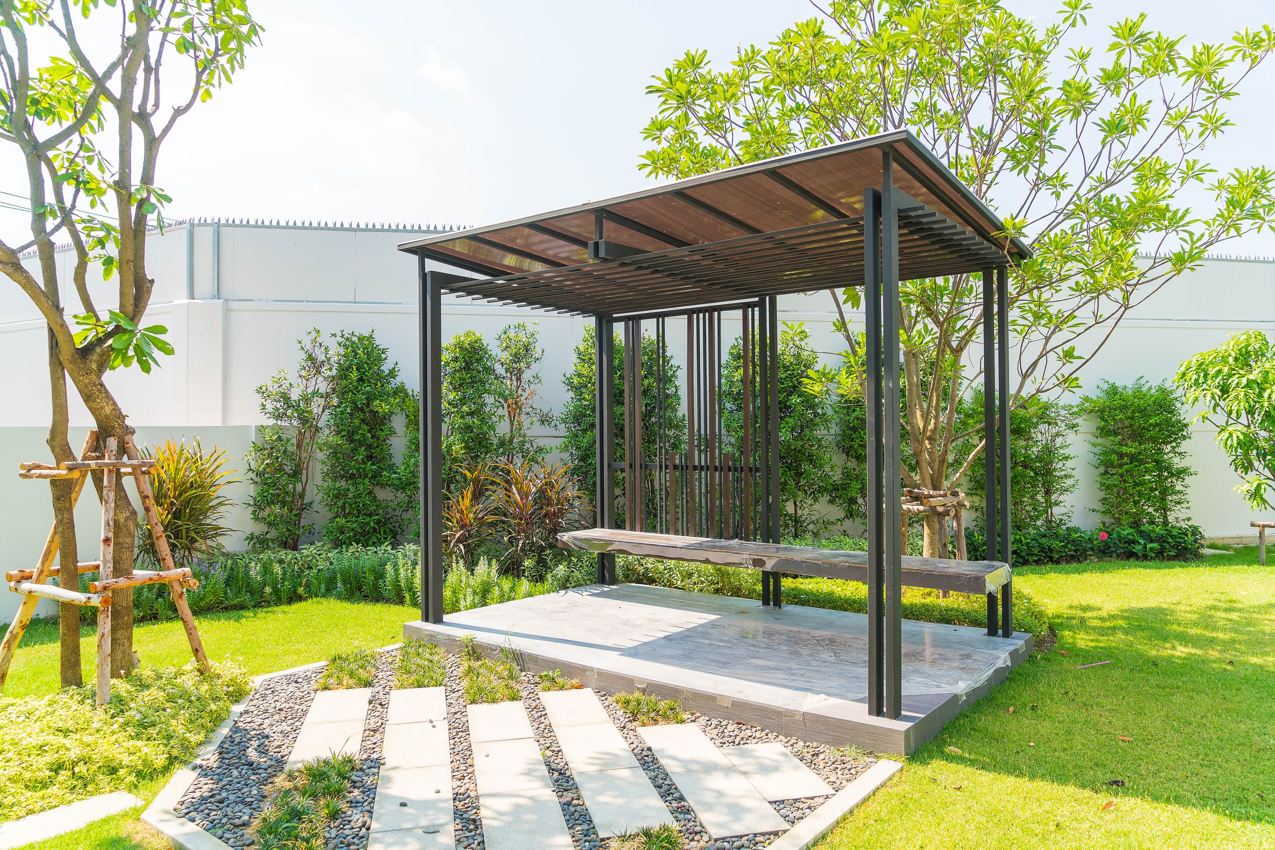 Modern black metal pergola with a slatted roof over a concrete patio, surrounded by a green lawn, hedges, trees, and garden beds.