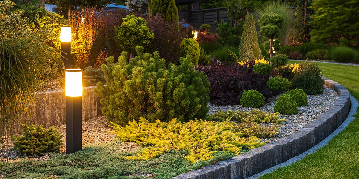 Warm outdoor pathway lights illuminate lush green shrubs and stone garden borders in a beautifully landscaped backyard setting.