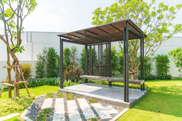 Modern black metal pergola with a slatted roof over a concrete patio, surrounded by a green lawn, hedges, trees, and garden beds.