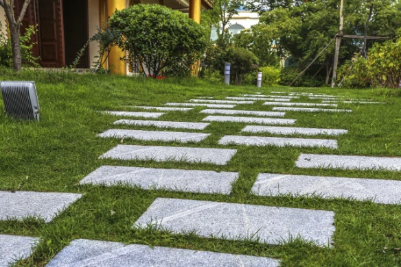 Rectangular gray stepping stones create a walkway across a green lawn toward a residential entrance, with low-profile path lights along the edge.