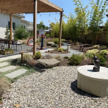 Backyard landscape with decorative river rocks, natural boulders, ornamental grasses, stepping stones, a fire feature, and a wooden pergola