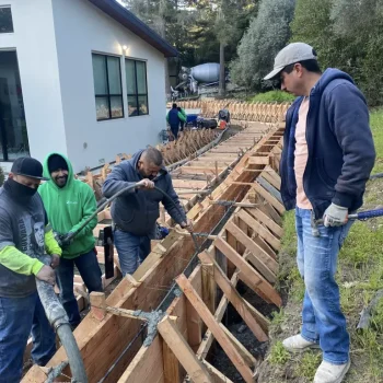 Landscape crew pouring concrete into wood retaining wall formwork with a mixer truck.