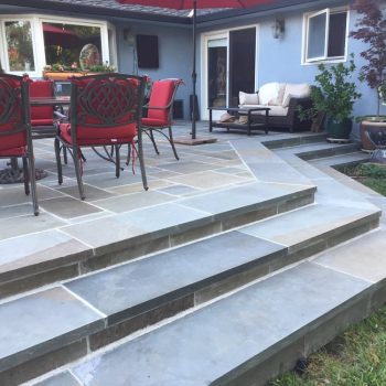 Commercial patio and walkway area with natural stone pavers, multi-tiered stairs, outdoor seating, dining furniture, and landscaped surroundings.