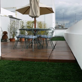 Rooftop terrace with a polished wooden deck, green turf sections, metal dining table, striped chairs, and a beige patio umbrella.